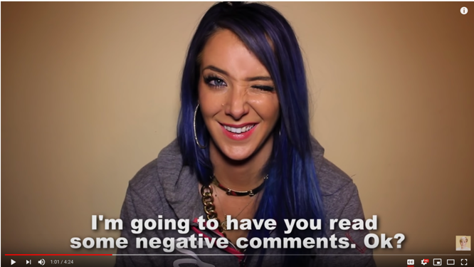 Jenna Mourey smiles and winks uncomfortably in response to Hannah Hart’s statement, captioned in white text at the bottom of the screen, “I’m going to have you read some negative comments. Ok?”