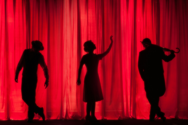 Image of three silhouetted figures performing against illuminated red stage curtains