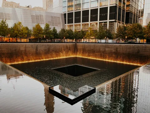 One of the 9/11 Memorial reflecting pools in New York City, with water cascading down its bronze-lit walls into a central void, surrounded by trees and modern buildings at dusk.