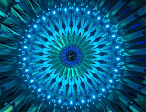 A radial kaleidoscopic pattern in shades of blue and teal, with concentric rings of pointed geometric shapes and glowing light points emanating from a central core.