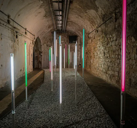 Colorful illuminated LED tubes standing upright on a gravel path inside a weathered stone tunnel or vaulted corridor, creating a light installation in a historic space.