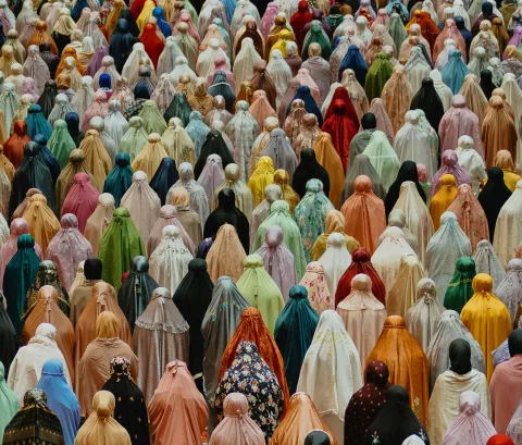An overhead view of a large gathering of women in prayer, wearing colorful headscarves and garments in a wide spectrum of pastel and vibrant hues.