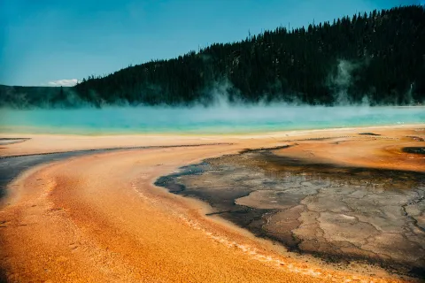 A steaming hot spring with vivid bands of orange, yellow, and turquoise water, with forested hills in the background.