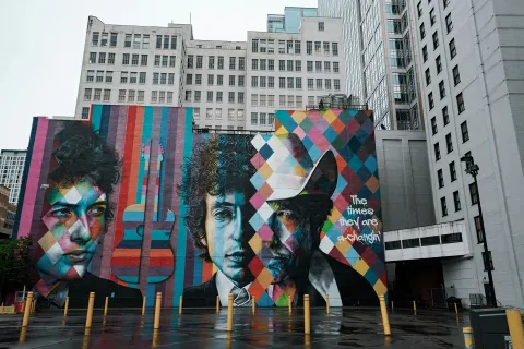 Colorful mural of three Bob Dylan faces painted on the side of a city building with the phrase, "The times they are a-changin'" painted to the right of them.