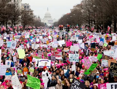 Massive crowd of demonstrators carrying colorful signs and wearing pink hats fills a broad avenue stretching toward the U.S. Capitol building on an overcast day.