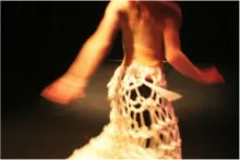 A motion-blurred dancer mid-movement wearing a white open-weave skeletal skirt or cage structure, photographed from behind on a dimly lit stage.