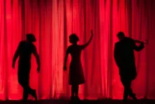 Image of three silhouetted figures performing against illuminated red stage curtains