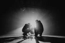 Two silhouetted figures crouch beside a film camera on a dimly lit set, backlit by a single dramatic beam of light cutting through haze.