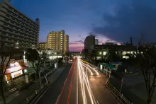 A long-exposure night shot captures streaking car light trails along a wide urban road in a city, set against a dramatic purple and pink twilight sky.