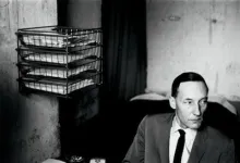 Black-and-white photo of a man in suit beside wall-mounted paper trays in sparse office setting.