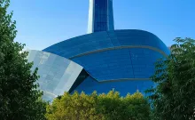 The Canadian Museum for Human Rights with sweeping curved blue glass facades and a tall glass tower rising from the center, framed by green trees under a clear blue sky.