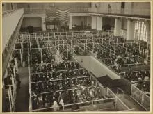 Black-and-white photo of large crowded hall with partitioned sections and people seated in organized rows.
