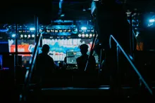 Silhouetted figures at a production control station with glowing monitors, backstage at a live event with colorful stage lighting in the background.