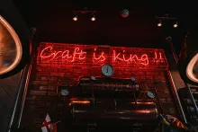 Red neon sign reading "Craft is King!!" mounted on a brick wall above copper brewing equipment with gauges and pipes in a dimly lit craft brewery.