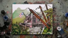 A painting on a work surface depicting a damaged house with fallen power line poles, a Puerto Rican flag, and overgrown vegetation, surrounded by ink and paint bottles.