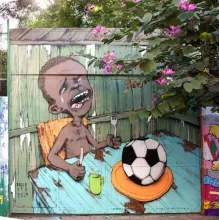 Street mural depicting a crying, emaciated child holding a fork over a plate with a soccer ball on it, with a small green cup beside it, signed "Paulo Ito 2014," partially framed by flowering branches.