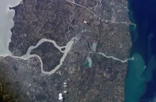 A sediment-laden river winds through a flat urban landscape before emptying into water.