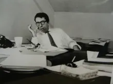 A black-and-white photograph of a man in glasses sitting at a desk and appearing mid-conversation
