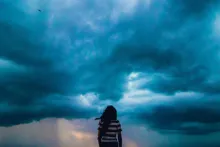 A small child, with their back to the camera, stares at a sky filled with storm clouds.