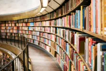 A curved library aisle lined with tall shelves filled with colorful books stretches into the distance under soft overhead lighting.