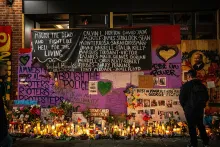 A nighttime memorial wall covered in handwritten messages, names of victims, photos, flowers, and lit candles, with text including "Mourn the Dead and Fight Like Hell for the Living" and "Rest in Power."